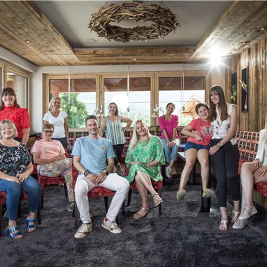 Eine Gruppe von Frauen und einem Mann sitzt in einem gemütlichen Raum mit Holzinterieur. Der Raum hat große Fenster, die viel Licht hereinlassen.