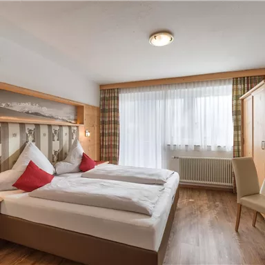 A cozy hotel room with two single beds and red pillows. Large windows provide plenty of daylight and a pleasant atmosphere.