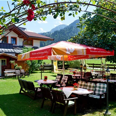 An inviting outdoor restaurant with tables under a sunshade. Surrounded by green trees and a beautiful mountain landscape.