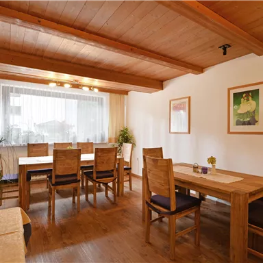 A cozy room with a wooden table and chairs. Large windows let in plenty of light, and plants create a pleasant atmosphere.