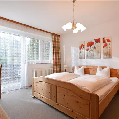 A cozy bedroom with a double bed and wooden furniture. Bright windows with curtains and a view of a balcony create a friendly atmosphere.