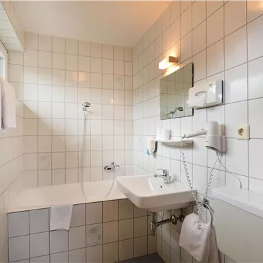 A modern bathroom with white tiles, a bathtub, and a sink. The shower is equipped with a handheld showerhead, and there are towels on the wall.