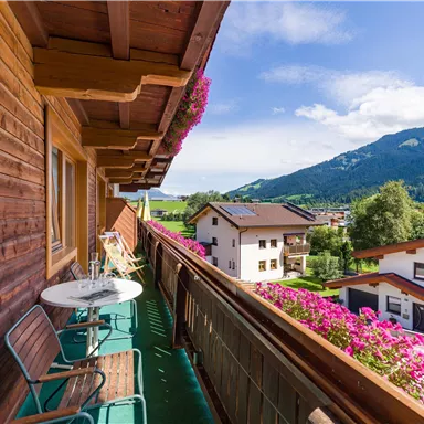 Ein Balkon mit einem Tisch und Stühlen, umgeben von bunten Blumen. Im Hintergrund sind grüne Berge und ein blauer Himmel zu sehen.