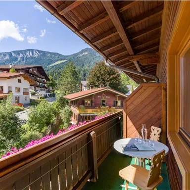Ein sonniger Balkon mit einem kleinen Tisch und Stühlen, umgeben von grünen Bäumen. Im Hintergrund sind Berge und traditionelle alpine Häuser zu sehen.