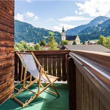 Ein Balkon mit einem Liegestuhl und Blick auf grüne Berge und das Dorf. Im Hintergrund ist eine Kirche sichtbar, umgeben von einer malerischen Landschaft.