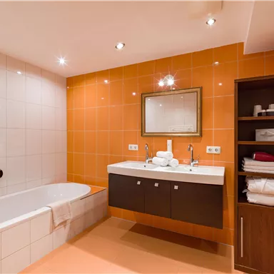 Das Badezimmer hat eine moderne Ausstattung mit einer Badewanne und einem eleganten Waschbecken. Die Wände sind in warmen Orangetönen gehalten, was eine einladende Atmosphäre schafft.