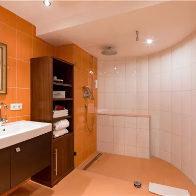 Ein modernes Badezimmer mit orangefarbenen Fliesen und einer eleganten Dusche. Die Einrichtung umfasst einen Waschtisch und einen Holzschrank mit Handtüchern.