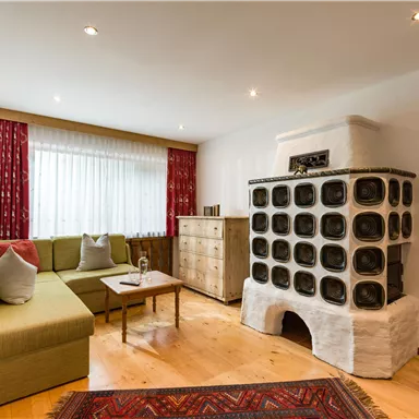 Ein gemütliches Wohnzimmer mit einer grünen Couch und einem traditionellen Ofen. Große Fenster lassen viel Tageslicht herein und die Dekoration ist einfach und einladend.