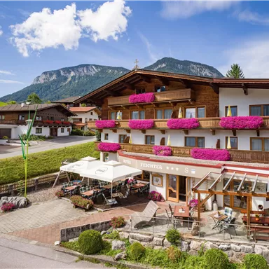 Ein charmantes alpenländisches Hotel mit bunten Blumenbalkonen. Im Hintergrund sind beeindruckende Berge und ein klarer Himmel zu sehen.