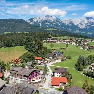 Eine malerische Landschaft mit charmanten Häusern und grünen Wiesen. Im Hintergrund erheben sich hohe Berge unter einem klaren blauen Himmel.