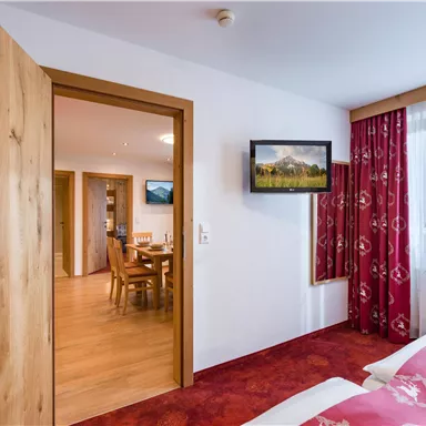 Ein gemütliches Schlafzimmer mit einem Fernseher und roten Vorhängen. Im Hintergrund sind ein Esstisch und weitere Räume zu sehen.