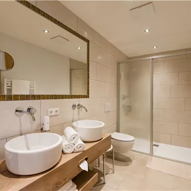 Ein modernes Badezimmer mit zwei Waschbecken und einer Dusche. Helle Fliesen und stilvolle Beleuchtung sorgen für eine angenehme Atmosphäre.