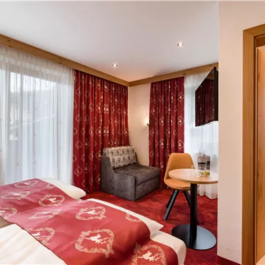 Ein gemütliches Hotelzimmer mit einem Doppelbett, einem Sitzbereich und großen Fenstern. Die Wände sind mit roten Vorhängen dekoriert, und es gibt einen kleinen Tisch.