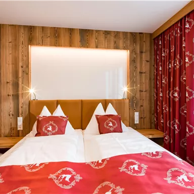 Ein gemütliches Schlafzimmer mit zwei Betten und roten Bettüberwürfen. Die Wände sind aus Holz, und es gibt helle Vorhänge.