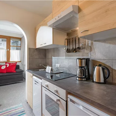 Eine moderne Küche mit Holz- und grauen Steinoberflächen. Im Hintergrund ist ein gemütliches Wohnzimmer mit einem Sofa und einem bunten Teppich sichtbar.