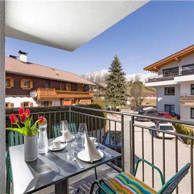 Ein schöner Balkon mit einem Tisch, auf dem Blumen und Gläser stehen. Im Hintergrund sind moderne und traditionelle Gebäude sowie Bäume zu sehen.