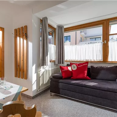 Ein helles Wohnzimmer mit großen Fenstern und einem gemütlichen Sofa. Ein Essbereich mit einem Tisch und frischen Blumen ergänzt den Raum.