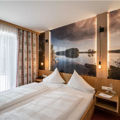 A modern bedroom with two beds and a beautiful wallpaper that depicts a landscape by the water. Large windows provide plenty of daylight.