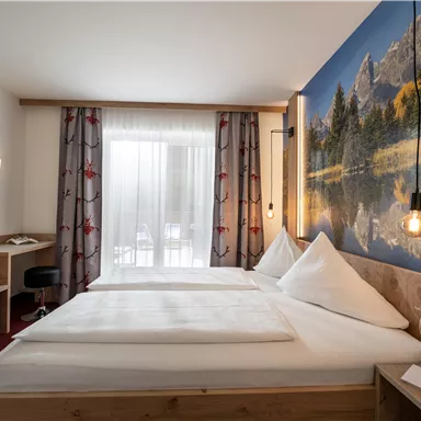 A cozy room with two beds and wooden decor. On the wall, there is a large landscape painting and there is plenty of daylight coming through the windows.
