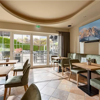 An inviting café with modern tables and chairs. Large windows and a television display a beautiful mountain landscape.