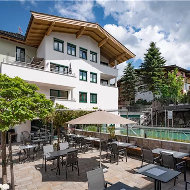 Ein modernes Gebäude mit Außenbereich und einem Pool ist zu sehen. Die Terrasse ist mit Tischen und Stühlen ausgestattet, umgeben von Bäumen und schönem Wetter.