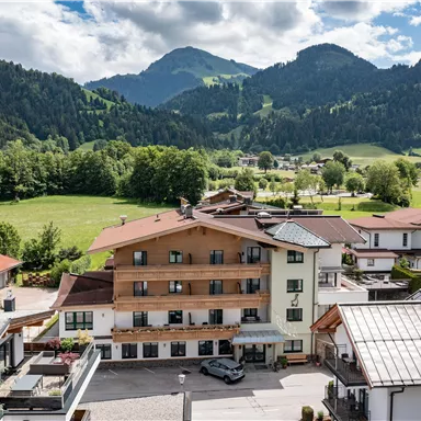 Ein malerisches Bergdorf mit modernen Häusern und vielen Bäumen. Im Hintergrund erheben sich beeindruckende Berge unter einem teilweise bewölkten Himmel.