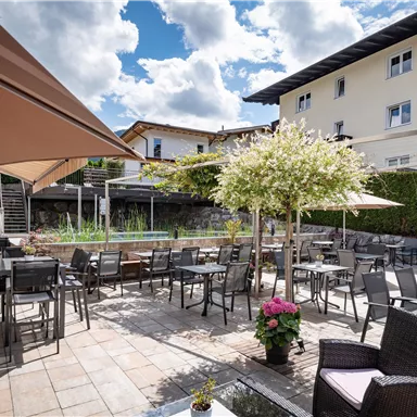 Eine einladende Terrasse mit Tischen und Stühlen unter Sonnenschirmen. Umgeben von Pflanzen und einem klaren Himmel.