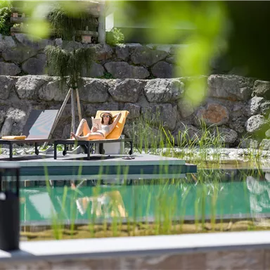 Eine entspannte Person liegt auf einer Liege am Wasser. Umgeben von grünen Pflanzen und Steinen, strahlt der Ort Ruhe und Erholung aus.