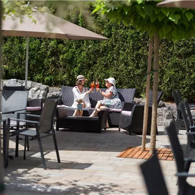 Zwei Frauen sitzen entspannt in einem Gartenbereich und genießen Drinks. Umgeben von Sonnenschirmen und Bäumen wirkt die Atmosphäre freundlich und einladend.