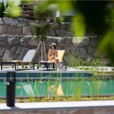 Eine Frau sitzt entspannt am Pool auf einer Liege. Umgeben von einem schönen Landschaftsdesign und klarem Wasser genießt sie die Ruhe.