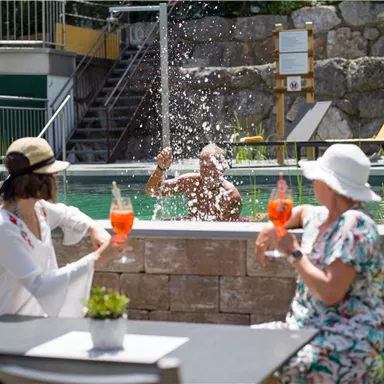Eine entspannte Atmosphäre am Pool mit zwei Frauen, die Getränke halten. Im Hintergrund plantscht ein Mann im Wasser.