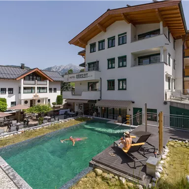 Ein modernes Hotel mit einer großen Terrasse und einem schönen Schwimmbecken. Im Hintergrund sind Berge und ein klarer blauer Himmel zu sehen.