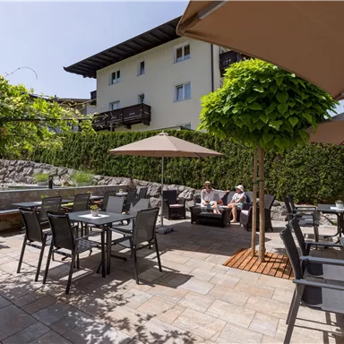 Ein gemütlicher Außenbereich mit Tischen und Stühlen unter Sonnenschirmen. Im Hintergrund sind grüne Sträucher und ein Gebäude sichtbar.