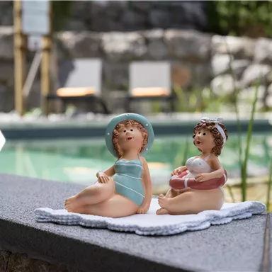 Zwei dekorative Figuren von Kindern am Poolrand. Eines sitzt in einem Badeanzug, während das andere ein Schwimmring hält.