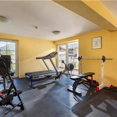 A modern fitness room with various equipment, including a treadmill and dumbbells. The walls are painted yellow and there are large windows with natural light.