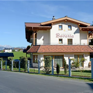 Ein charmantes Gebäude mit der Aufschrift „Bavaria“ in einer ländlichen Umgebung. Im Hintergrund ist eine Kirche und eine grüne Landschaft zu sehen.