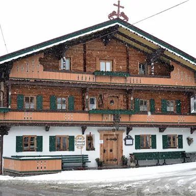 Ein traditionelles, mehrstöckiges Chalet im Alpenstil, umgeben von schneebedeckten Landschaften. Die Fassade ist aus Holz mit grünen Fenstern und Balkonen.
