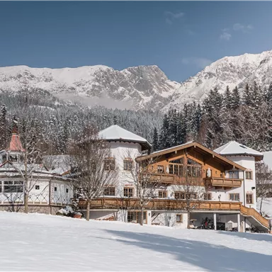 A charming house in a snowy landscape, surrounded by mountains. The sky is clear and there are many trees nearby.