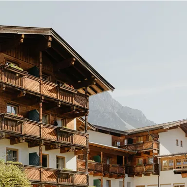 Ein charmantes Gebäude im alpinen Stil mit Balkonen und Holzverkleidung. Im Hintergrund sind Berge zu sehen, die eine malerische Landschaft bieten.