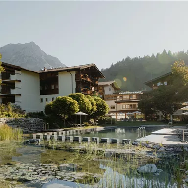 Ein malerisches Hotel in den Bergen, umgeben von Bäumen und Sträuchern. Im Vordergrund ist ein ruhiger Teich mit modernen Liegen und Sonnenschirmen.