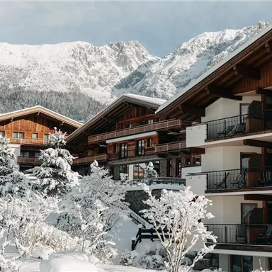 Eine malerische Winterlandschaft mit schneebedeckten Hütten und hohen Bergen im Hintergrund. Die Häuser haben holzverkleidete Fassaden und sind von schneebedeckten Bäumen umgeben.