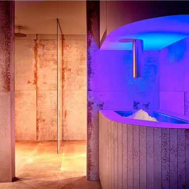 A modern wellness area with soft lights in shades of purple. The walls are made of concrete, and an elegant, curved counter is visible.