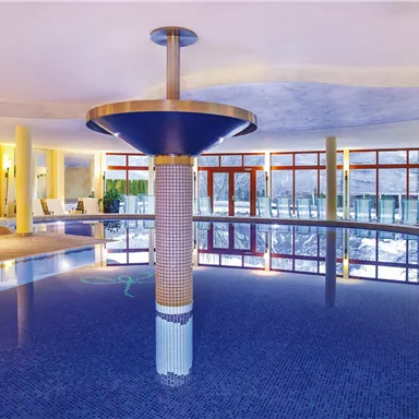 An elegant indoor pool with blue water and an open, bright atmosphere. Green plants and comfortable loungers create a relaxing environment.