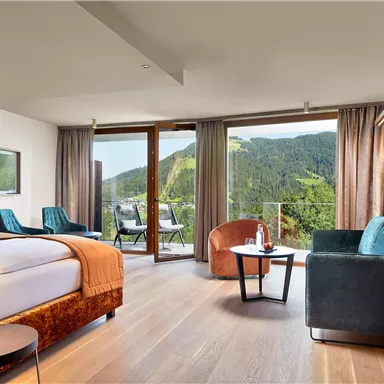 A modern hotel room with large windows and a view of the mountains. It is stylishly furnished with a bed, sofa, and a small table.