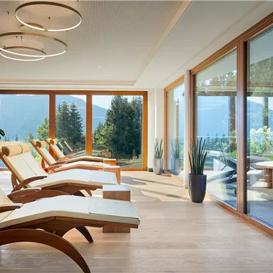 A modern wellness area with wooden loungers and large windows. In the background, green mountains can be seen.