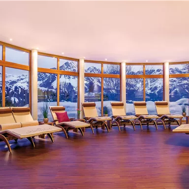 A stylish relaxation room with loungers and large windows. In the background, snow-covered mountains and a gentle evening sky can be seen.