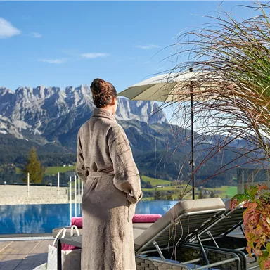 A person in a bathrobe stands next to a pool, looking at the mountains. The sky is clear, and the surroundings are calm and inviting.