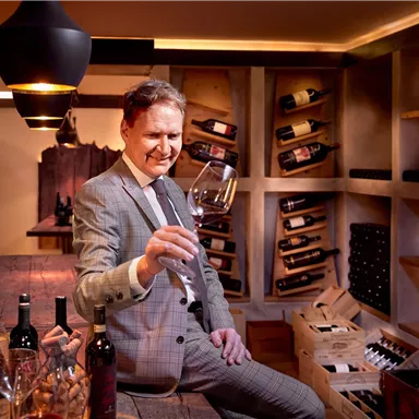A man in an elegant suit holds a wine glass in a stylish wine cellar setting. In the background, wine bottles and shelves are visible.