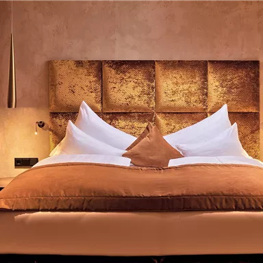 An elegant bedroom with a large bed and stylish pillows. The wall behind the bed is painted in warm, golden tones.