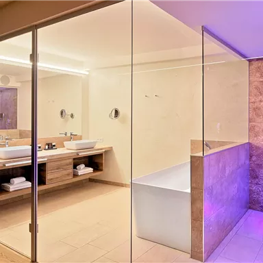 A modern bathroom with an elegant glass partition. It features a bathtub and two sinks, surrounded by warm light.